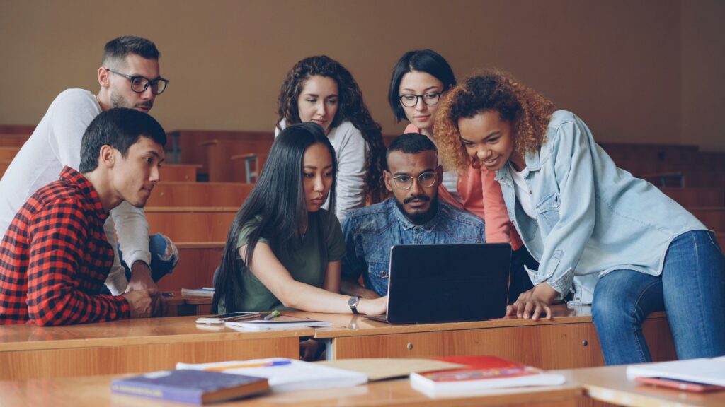 A diverse group of young students leaning over a laptop together in a lecture hall, discussing what is on the screen